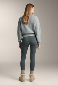 Szürke rövid pulóver széles ujjal, párosítva bordázott zöldeskék leggingsekkel. A modellen bézs bokacsizmák vannak vastag kötött zoknikkal.