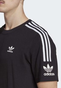 Muž nosící černé tričko Adidas s bílým designem tří pruhů na ramenou a logem Adidas na hrudi a rukávu.