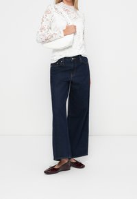 Vit långärmad spetsblus med blommönster, mörkblå vidbyxade jeans och mörkröda lackballerinas. Handväskan är vit med en böjd design.