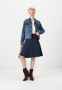Modrá džínsová bunda spárovaná s námorníckou sukňou s rozšírením. Modelka má na sebe hnedé členkové topánky a v ruke drží malú hnedú kabelku, na neutrálnom pozadí.