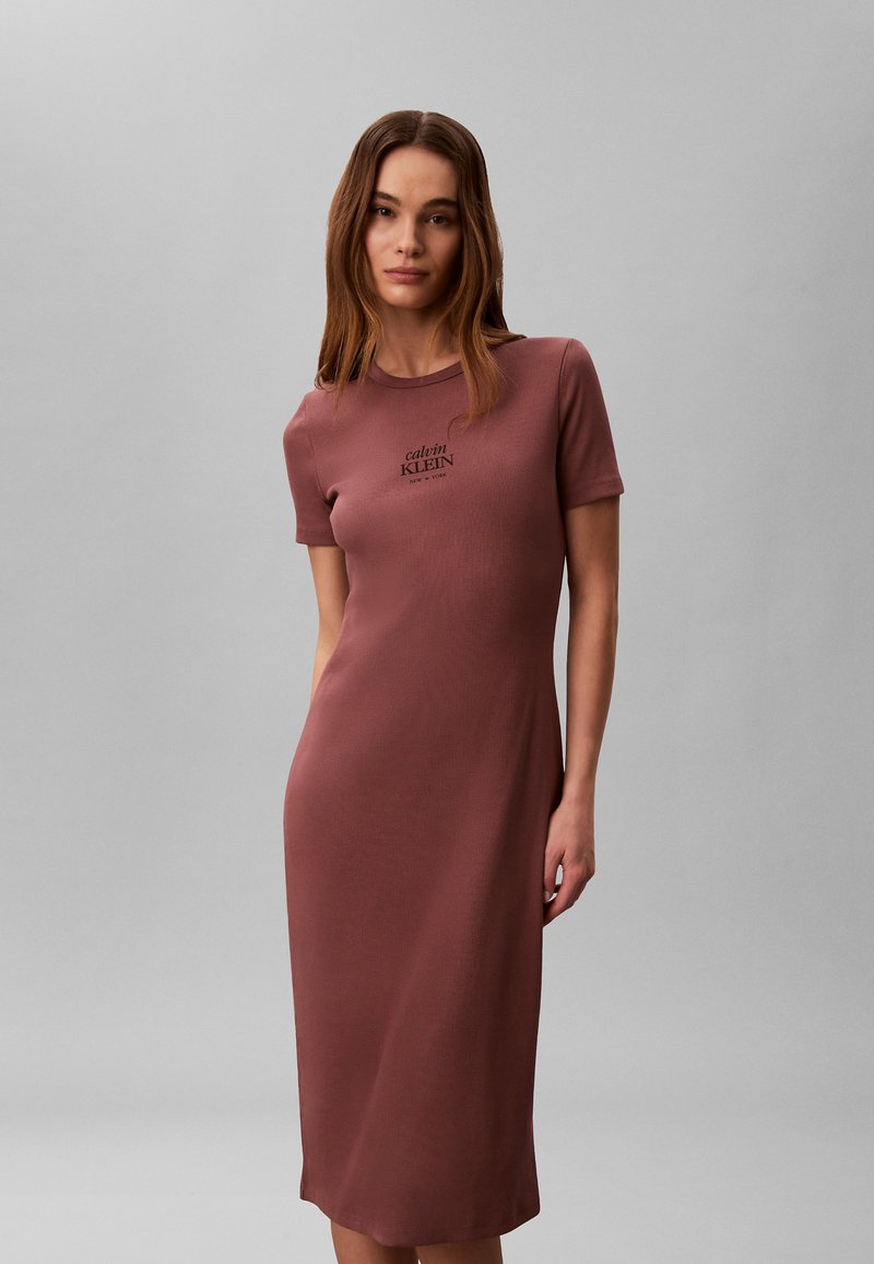 Vrouw met lang bruin haar, gekleed in een nauwsluitende, korte mouw, midi-jurk in stoffig roze met Calvin Klein-logo op de borst, voor een grijze achtergrond.