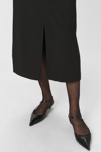 Czarna midi spódnica z przodu z rozcięciem, zestawiona z czarnymi balerinami na spiczastym nosku i przezroczystymi czarnymi rajstopami, prezentująca elegancki design i materiał.