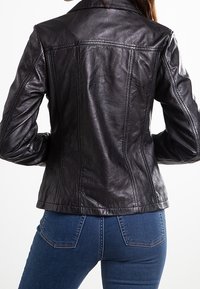 Sort læderjakke med en struktureret design, synlige sømme og en klassisk krave. Båret over blå jeans, som fremhæver en tætsiddende silhuet.