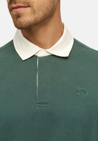 Groene katoenen polo met een witte kraag, voorzien van een knoopsluiting en een geborduurd logo met "EST. 1983" in het groen.