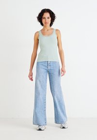 Top in cotone verde chiaro a coste con bordi ondulati abbinato a jeans blu chiaro a gamba larga. Modella che indossa sneaker grigie, in piedi contro uno sfondo bianco.