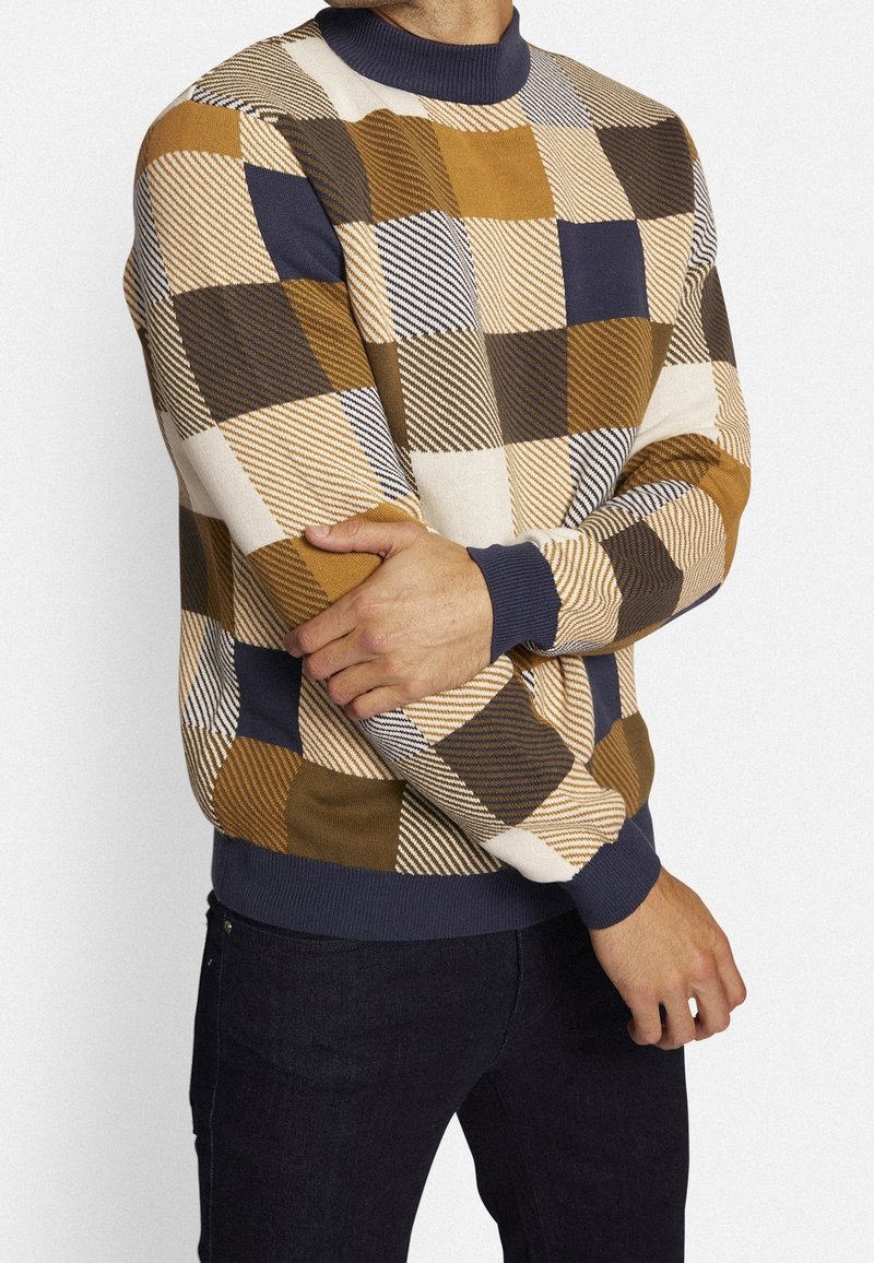 Homme portant un pull à carreaux multicolores en marron, beige, marine et blanc, associé à un pantalon foncé, debout les bras croisés.