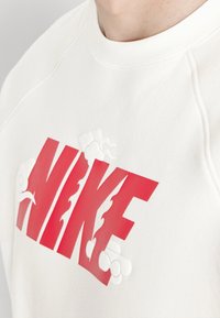 Vit sweatshirt med en stor röd "NIKE"-logotyp med flammande detaljer och molngrafik, tillverkad av mjukt tyg med ribbad halsringning.