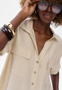 Beige button-up shirt with a collar, featuring two front pockets. Smooth texture, short rolled sleeves, and wooden buttons.