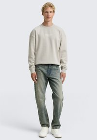 Sudadera gris claro con cuello redondo y corte relajado, combinada con jeans azules y zapatillas blancas. Presenta una marca mínima en el pecho.