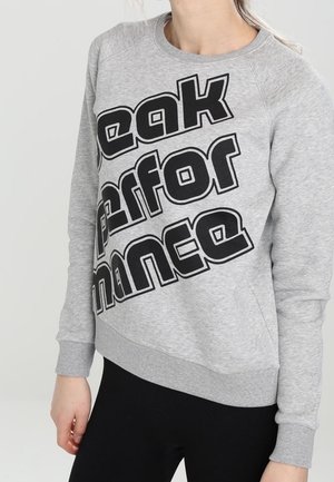 Osoba w jasnoszarej bluzie z dużym, czarnym, pogrubionym napisem „Peak Performance” drukowanym ukośnie na przodzie.