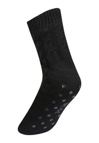 Calzino nero lavorato a maglia con motivo a treccia e punti in gomma antiscivolo sulla suola, su uno sfondo bianco.