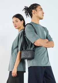 Black rectangular bag with embossed logo, detachable strap. Model wears a faded green oversized t-shirt. Simple design, minimalistic features.