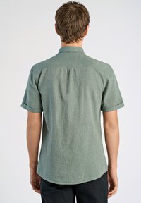 Camisa de manga corta con cuello en tejido entrelazado de color verde claro. Presenta un corte recto, una textura sutil y puños enrollados. Detalle de pliegue en la parte posterior visible.
