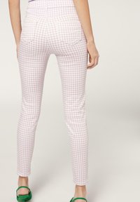 Mujer vistiendo pantalones de cuadros en tonos rosa y blanco con bolsillos en la parte trasera, combinados con sandalias de tacón verdes, de pie frente a un fondo neutro.