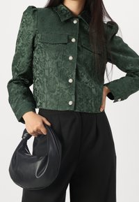 Grön texturerad jacka med krage, dekorativa framfickor och silverknappar. Kombinerad med en svart väska och högmidjade byxor.