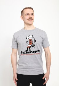 Camiseta de algodón gris con un dibujo de una salchicha en un gorro de chef, sosteniendo una espátula. El texto dice "¡Come Salchichas!" con un acento rojo.