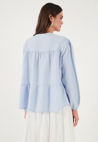 Blusa azzurro chiaro a maniche lunghe con vita arricciata, tessuto testurizzato e scollatura rotonda. L'orlo svasato e i polsini elasticizzati conferiscono forma.