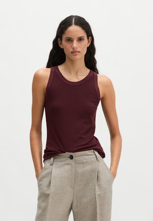 Femme aux longs cheveux foncés, portant un haut sans manches rouge foncé et un pantalon beige clair taille haute, les mains dans les poches.