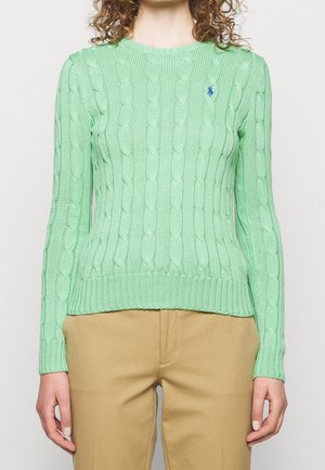 Femme portant un pull en maille torsadée vert clair et un pantalon beige, montrant le torse et les bras sur un fond uni.