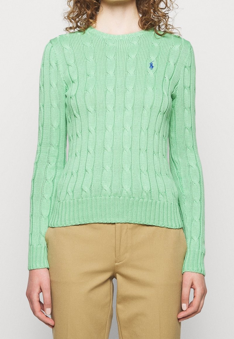Femme portant un pull en maille torsadée vert clair et un pantalon beige, montrant le torse et les bras sur un fond uni.