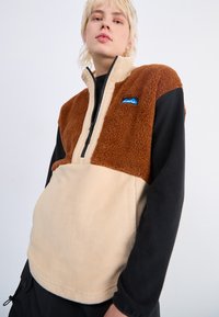 Sudadera de forro polar con diseño de media cremallera, que presenta secciones en marrón y beige y una manga larga negra. Incluye un detalle de logo azul en el pecho.
