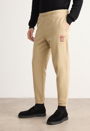 Beige Jogginghose mit elastischem Bund und Seitentaschen, mit rotem Lacoste-Logo. Weicher Stoff mit geradem Bein. Darunter schwarze Schuhe.