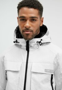 Chaqueta blanca impermeable con capucha, cremallera negra y detalles en negro, dos bolsillos delanteros y un parche de logo en el pecho. Textura suave.
