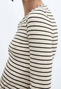 Nainen, jolla on päällään beige ribbipaita, jossa on vaakasuuntaisia mustia raidoja, kuvattuna sivulta yksinkertaista valkoista taustaa vasten.