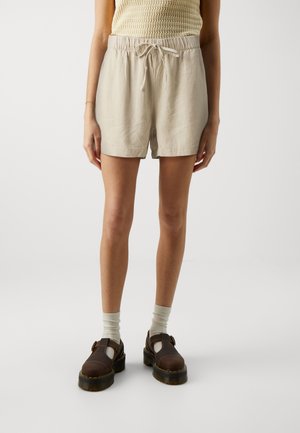 Pantaloni shorts in lino beige con vita elastica e coulisse. Indossati con scarpe marroni e calzini chiari.