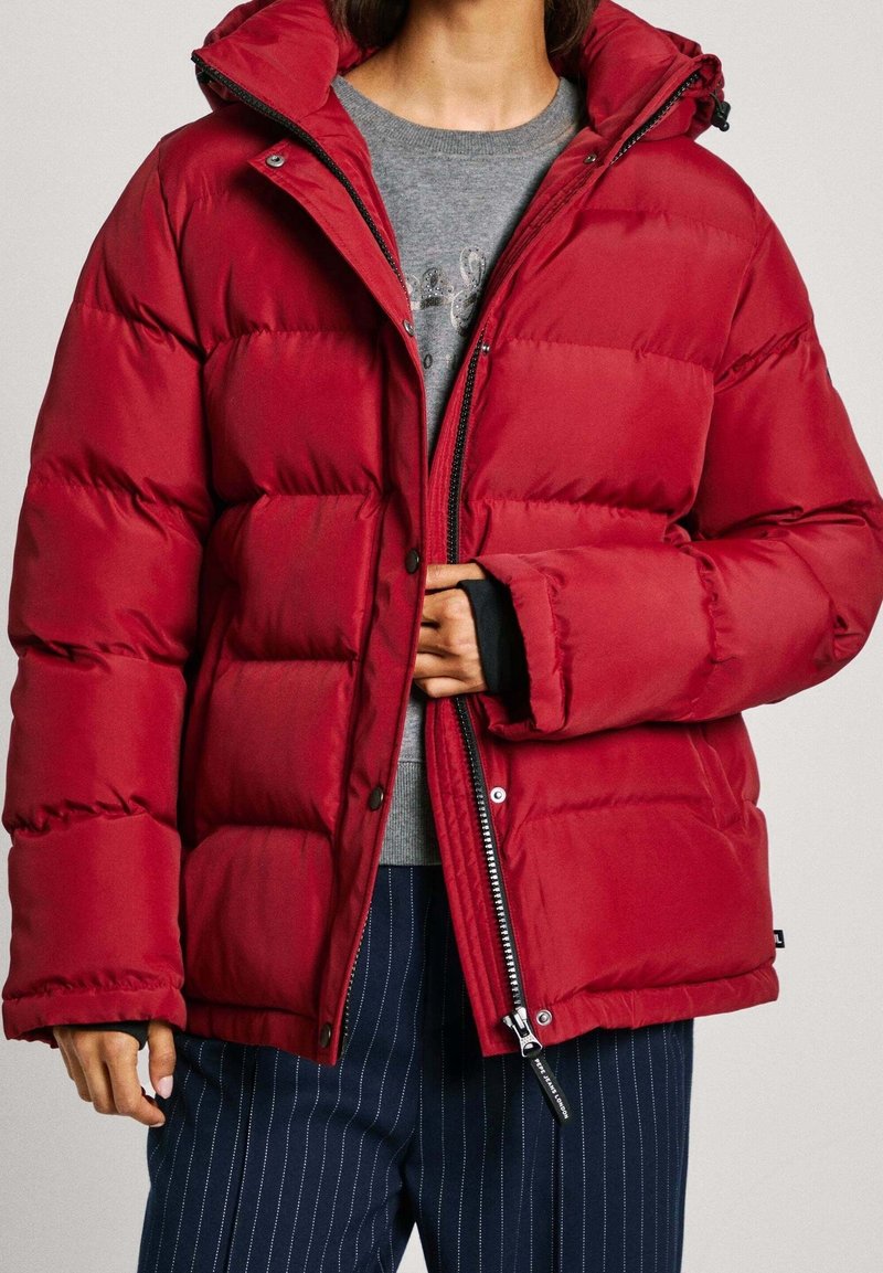 Röd puffjacka med huva, med ett quiltat mönster, dragkedja fram och tryckknappar, samt elastiska ärmslut; kombinerad med grå tröja och randiga byxor.