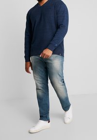 Navy knit sweater with a v-neck, paired with faded blue jeans and white sneakers featuring red accents. Texture is ribbed and smooth.