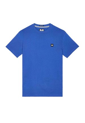 Mėlynas medvilninis marškinėlis su apvalia apykakle, trumpomis rankovėmis ir mažu juodu kvadratiniu ženkliuku krūtinėje. Lygi tekstūra ir laisvas kirpimas.