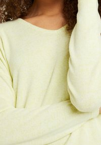 Jasnożółty sweter wykonany z miękkiego dzianinowego materiału, o luźnym kroju i szerokim dekolcie w kształcie litery V. Delikatna, nakrapiana faktura i długie rękawy.