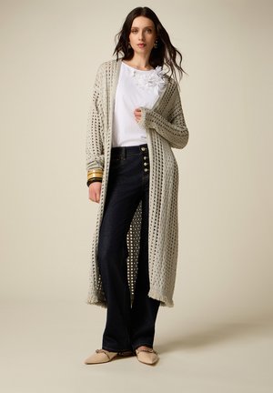 Kobieta w beżowym długim kardiganie z dzianiny, białej bluzce zdobionej kwiatami, ciemnych dżinsach na guziki, beżowych balerinach i złotych bransoletkach, stoi z rozpuszczonymi włosami.