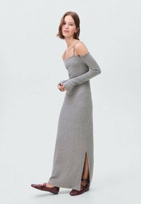 Abito maxi grigio a costine con maniche lunghe, design off-shoulder, spacco laterale, abbinato a scarpe flat burgundy. Texture liscia e silhouette aderente.