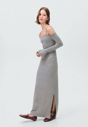 Robe maxi gris à côtes avec manches longues, design dénudé des épaules, fente latérale, associée à des chaussures plates bordeaux. Texture lisse et silhouette ajustée.