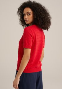 Punane lühikeste varrukatega sviiter, millel on tekstuuriga täppmustrid, ribaga mansetid ja istuv äär, koos tumesiniste pükstega.