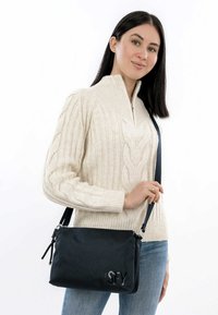 Borsa a tracolla nera in tessuto, con cerniera e un logo discreto. Modello che indossa un maglione a trecce di colore crema e jeans blu.