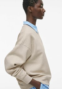 Beżowa bluza z oversize'owymi rękawami, z jasnoniebieską koszulą z kołnierzykiem pod spodem. Gładka tekstura, minimalistyczny design i luźny krój.