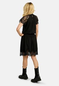 Sieviete ar blondiem matiem, valkā melnu ceļgalu garuma kleitu ar mežģīņu piedurknēm un apakšu, kopā ar melniem potīšu zābakiem, stāvot ar muguru pagriezta.