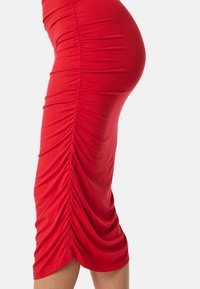 Robe ajustée rouge avec des détails froncés sur le côté, longueur genou et tissu lisse et extensible. Pas de matériels ou motifs visibles.