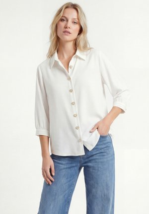 Vrouw draagt een witte blouse met knopen in hartvorm en blauwe jeans, poseert met één hand in haar zak tegen een effen achtergrond.