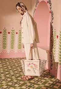 Sieviete valkā krēmkrāsas jaku un svītrainu galvas lakatu, rokās turot lielu rakstainu somu, fona ir ziedu un ģeometriska sienas gleznojuma motīvs.