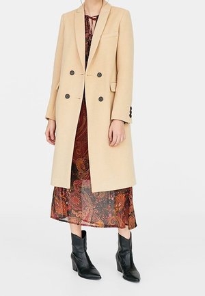 Beige dubbelbreasted jas over een lange, gemêleerde jurk, gecombineerd met zwarte enkellaarsjes met hak, gedragen door een staande persoon.