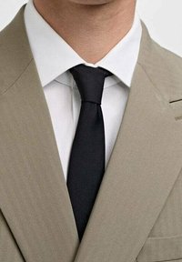 Uomo che indossa un giubbotto di abito beige, una camicia bianca e una cravatta nera ben annodata.