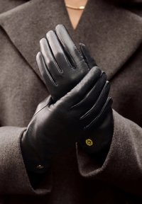 Gants en cuir noir avec une texture lisse, un design ajusté et de subtils détails de couture, portés sur un fond de tissu texturé sombre.