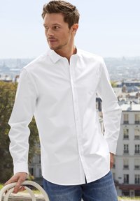 White long-sleeve shirt in cotton fabric with a button-down collar, paired with denim jeans. Features a straight-cut hem and minimal wrinkles.