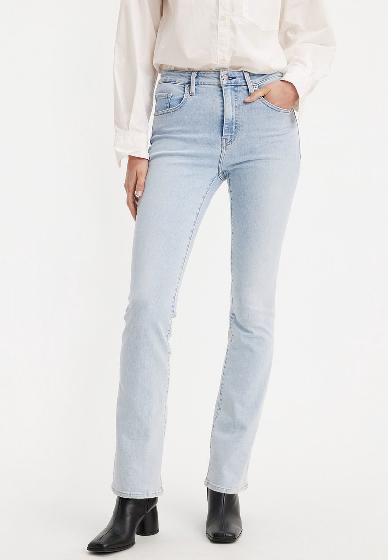 Personne portant un jean skinny taille haute bleu clair, une chemise blanche à manches longues et des bottines noires à talons, debout avec une main dans la poche.