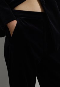 Schwarze Samt-Hose mit einer Seitentasche, im lockeren Schnitt und mit subtiler Glanzoptik. Der Stoff wirkt weich und glatt, was den Tragekomfort erhöht.