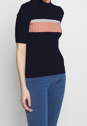 Frau trägt ein marineblaues, kurzärmeliges Ripptop mit weißen und pfirsichfarbenen horizontalen Streifen über der Brust, kombiniert mit blauen Jeans.